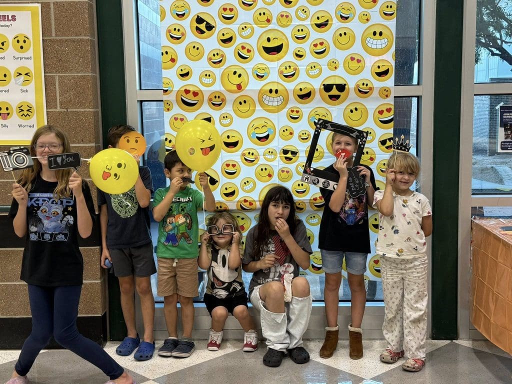 7 children pose in front of a poster with different emojis, holding various props.