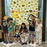 6 children pose in front of a poster with different emojis, holding various props.