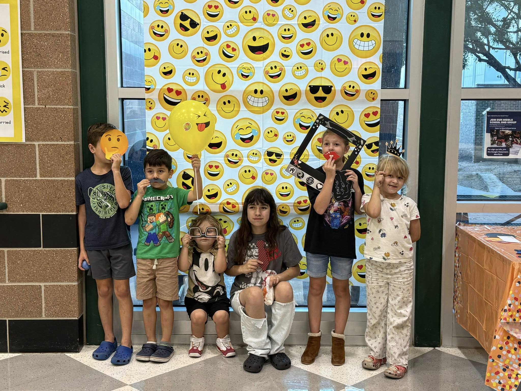 6 children pose in front of a poster with different emojis, holding various props.
