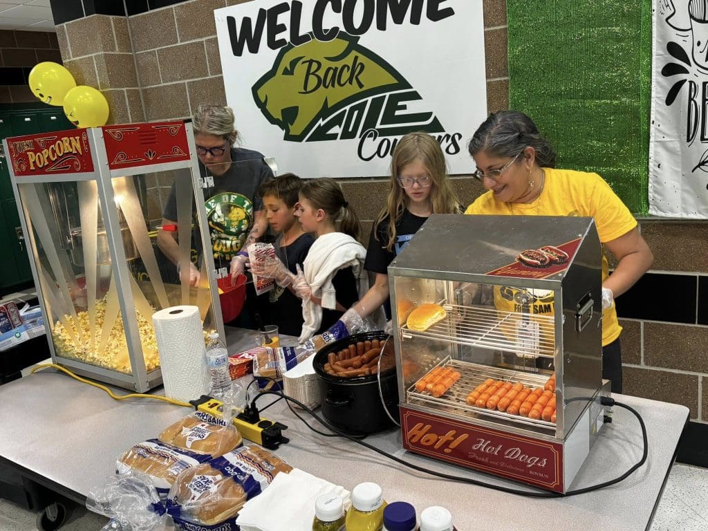 2 adults standing behind a table serve popcorn and hotdogs to 3 children.