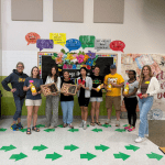 A group of 9 volunteers standing in front of a wall holding donuts and drinks.