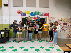 A group of 9 volunteers standing in front of a wall holding donuts and drinks.