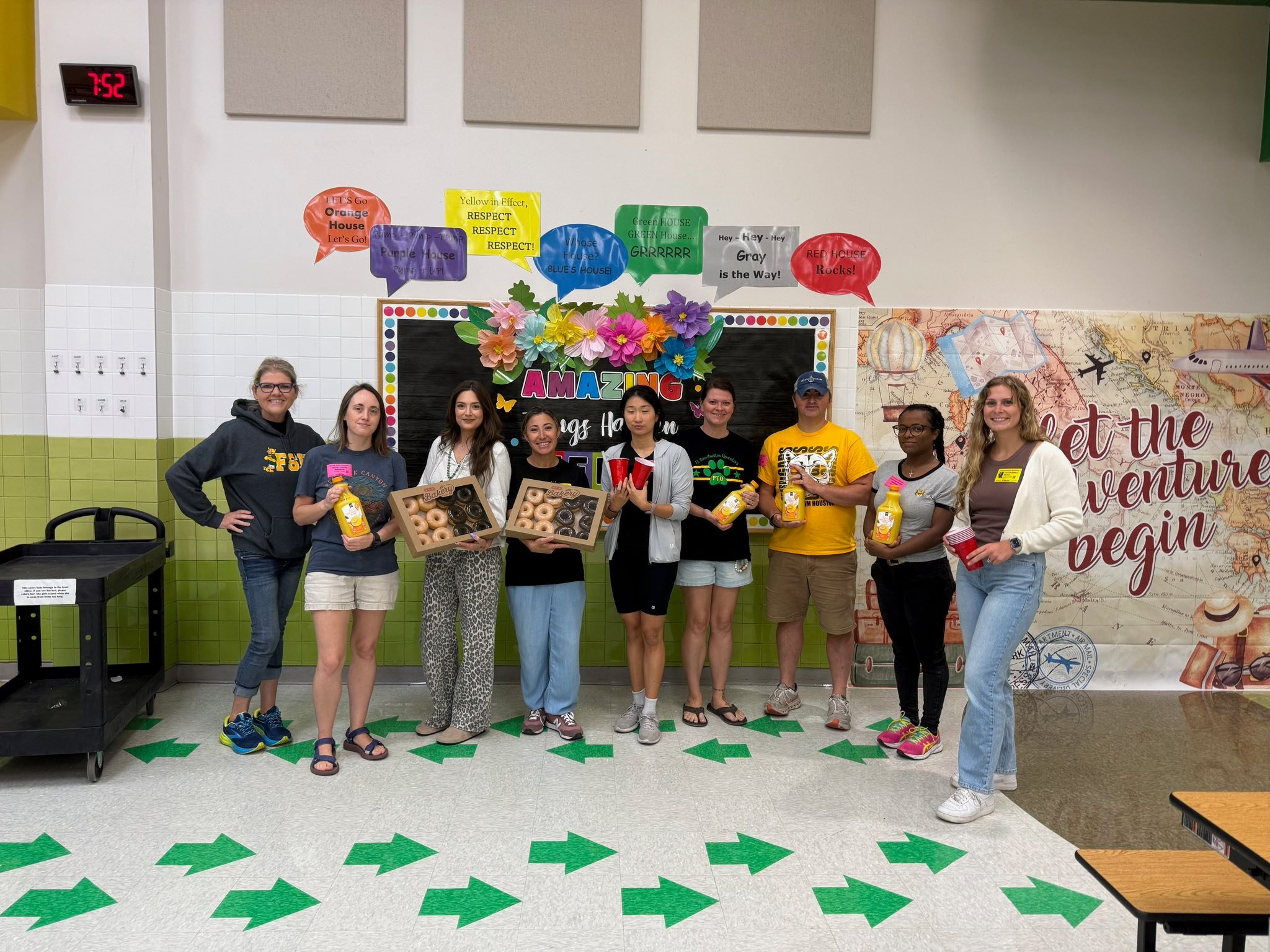 A group of 9 volunteers standing in front of a wall holding donuts and drinks.