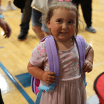 A little girl smiling for a photo, proudly wearing her new backpack.