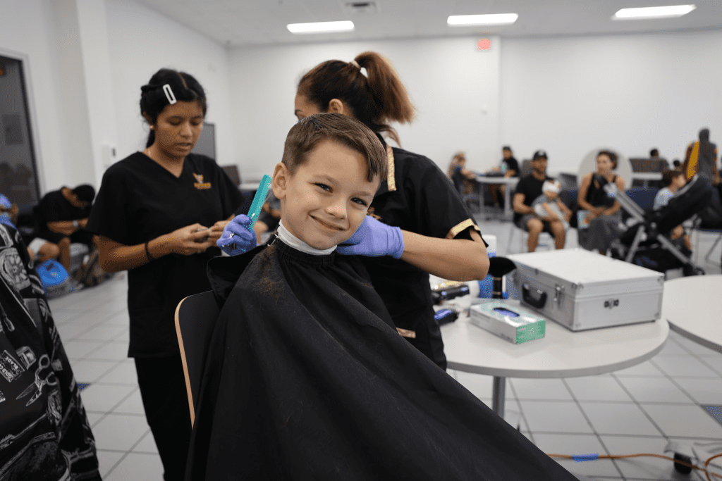A little boy sits in a salon chair, smiling at the camera while receiving a haircut.