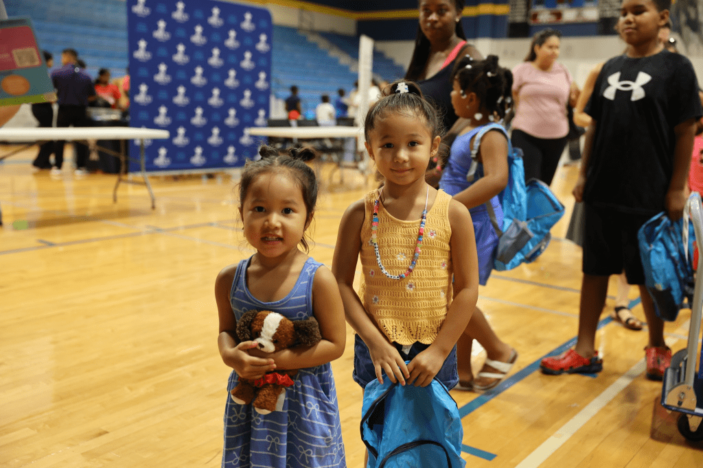 2 little girls pose for a photo, one holding a new backpack.