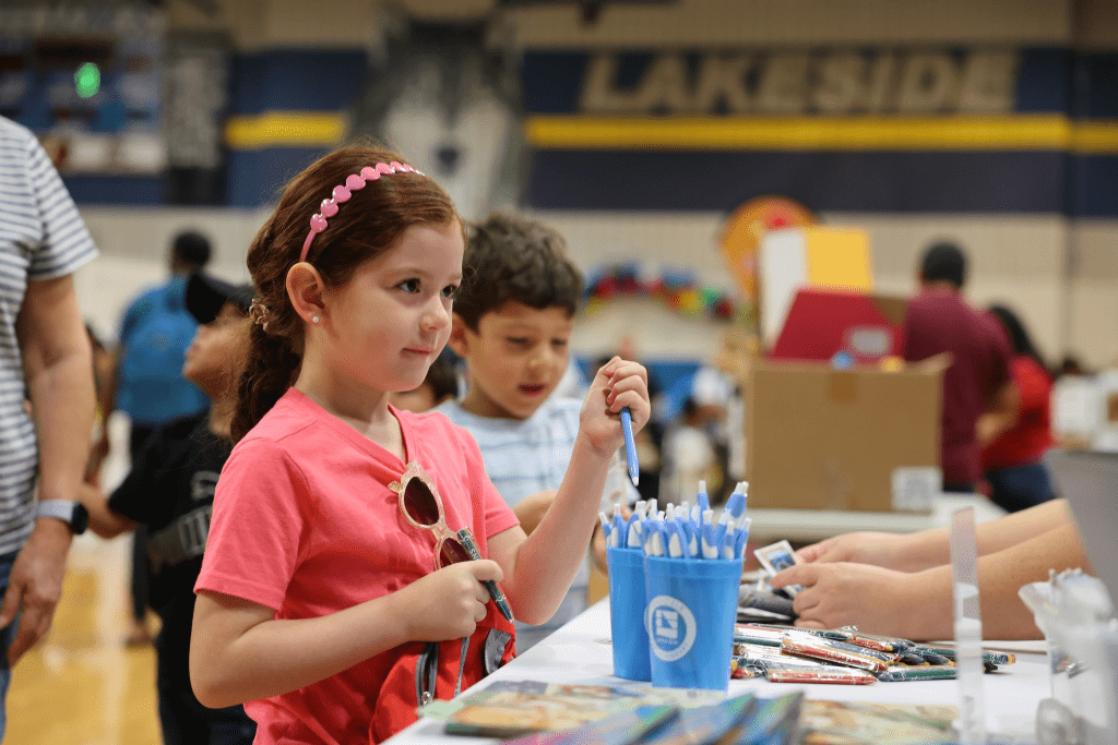 Students stand at an event table, one student holding her new backpack and grabbing a free pen.
