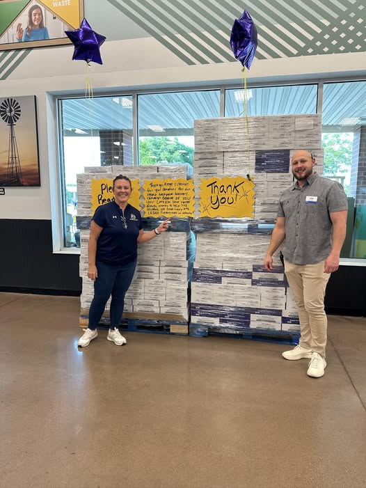 Two people stand in front of two pallets full of school supply kits.